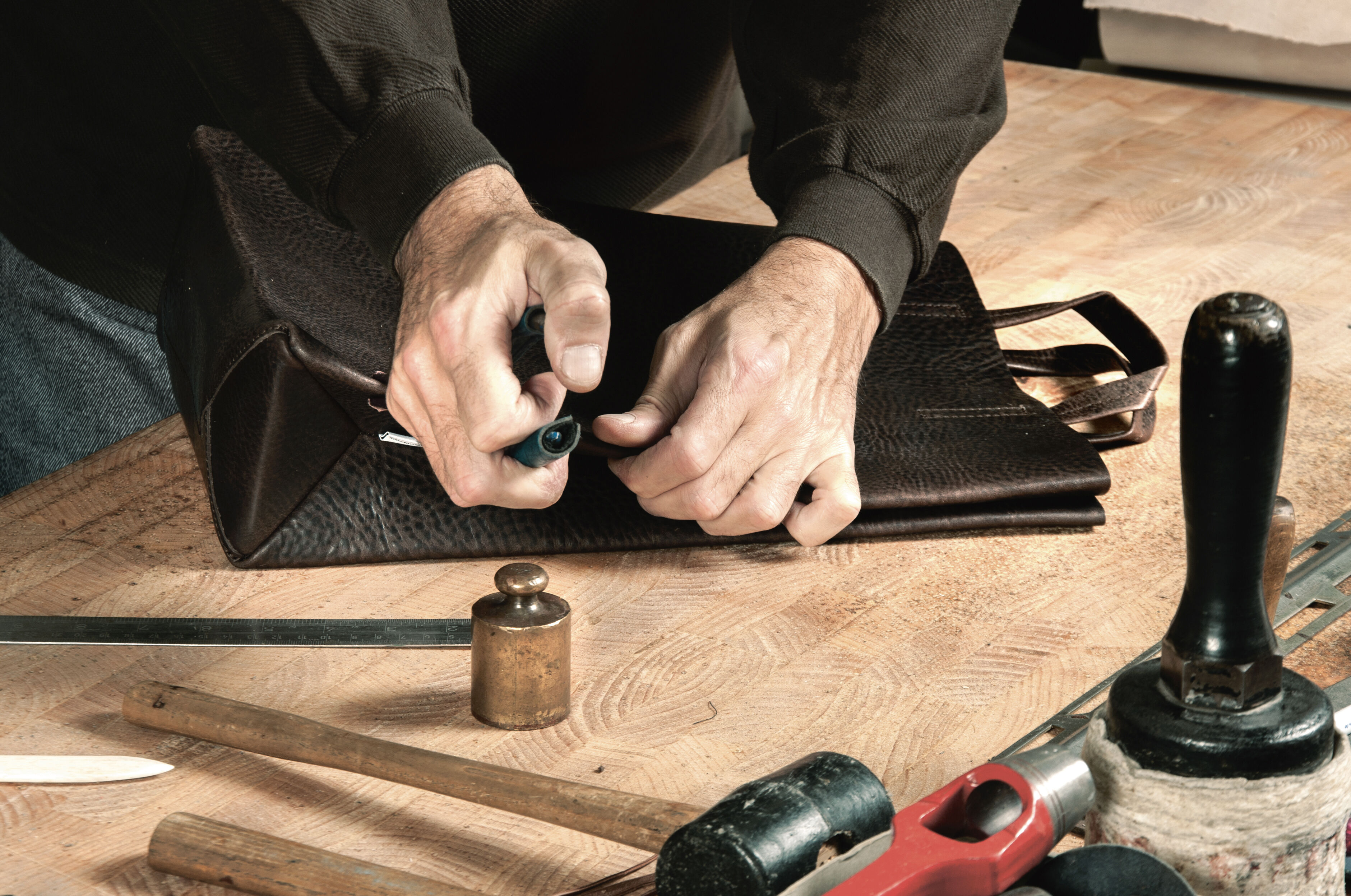 Hände falten eine schwarze Ledertasche auf einem Holztisch mit Werkzeugen und einer Metallgewicht